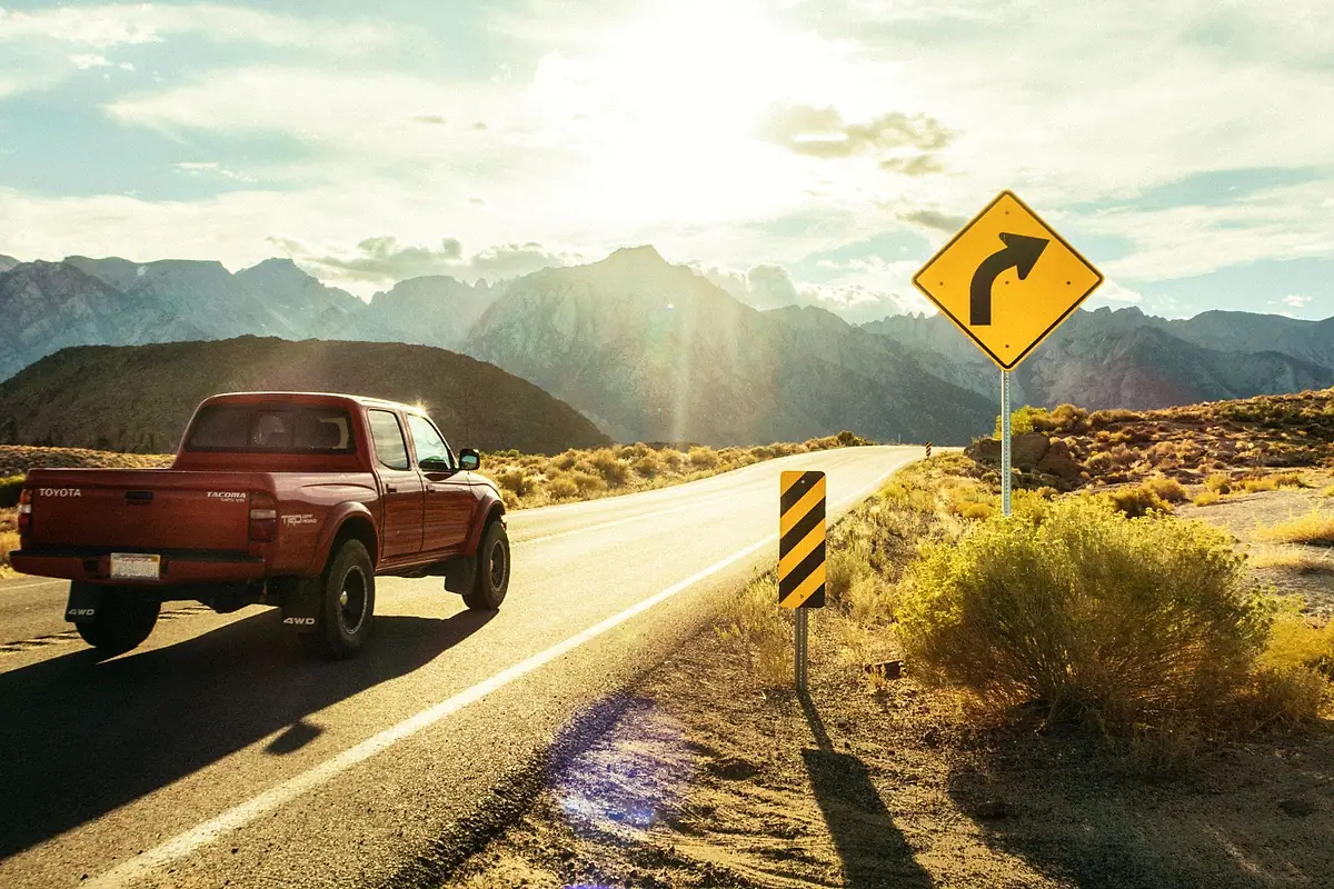 A red pickup truck on a two-lane desert highway, passing a yellow curve warning sign.